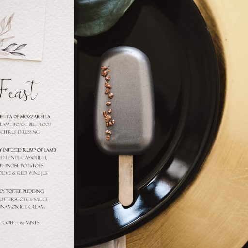 silver metallic cakesickle wedding favour with bronze crunch details on a black plate beside a wedding menu.