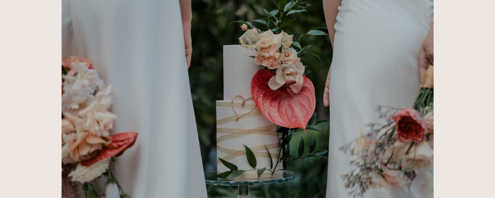 Striking two tier white wedding cake. Bottom tier has a sheer beige ribbon criss-crossed over the etirity, top thier is simle white fondant with a very large bright pink-red anthurium and contrasting smaller florals