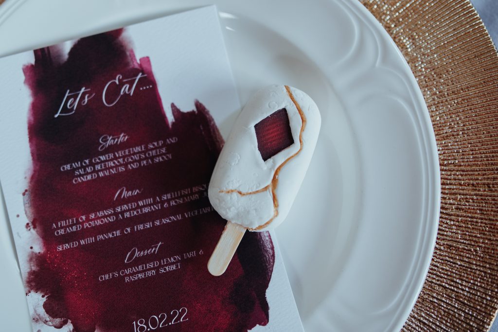 White cakesickle - looks like a mini magnum, but is filled with a coconut and lime cake with a lime buttercream. Covered in White chocolate. Robed in a very thin white fondant, kintsugi effect gold lines run throught and an edible red diamond applique. Fully edible