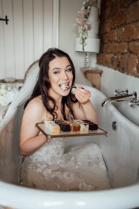 Bride sitting in a copper bathtub, in her wedding dress, with a plate of cake samples and eating a piece with a copper fork