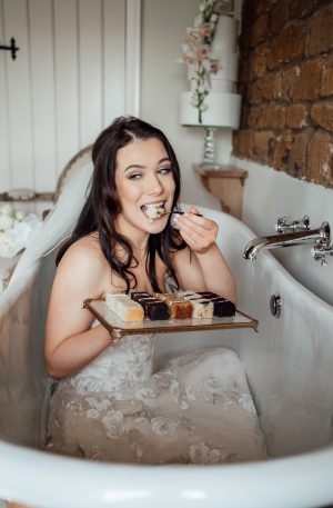 Bride sitting in a copper bathtub, in her wedding dress, with a plate of cake samples and eating a piece with a copper fork