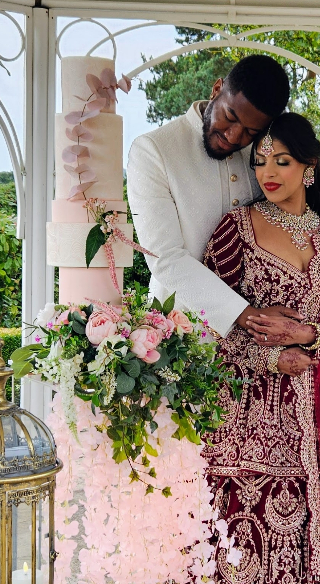Design-led wedding cake featuring peach and blush tones, sculptural tiers and cascading florals, thoughtfully styled within a contemporary outdoor pagoda wedding setting in Northamptonshire, alongside the bride and groom