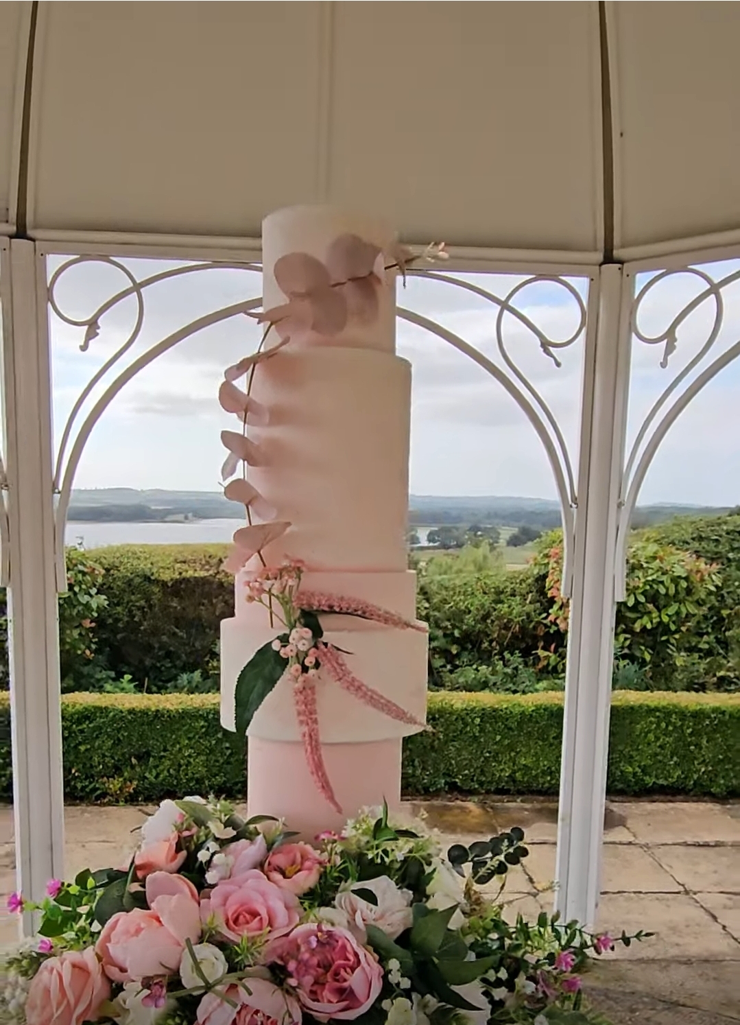 Blush and peach tone 5 tier wedding cake set in an outdoor setting on a floral plinth in a pagoda