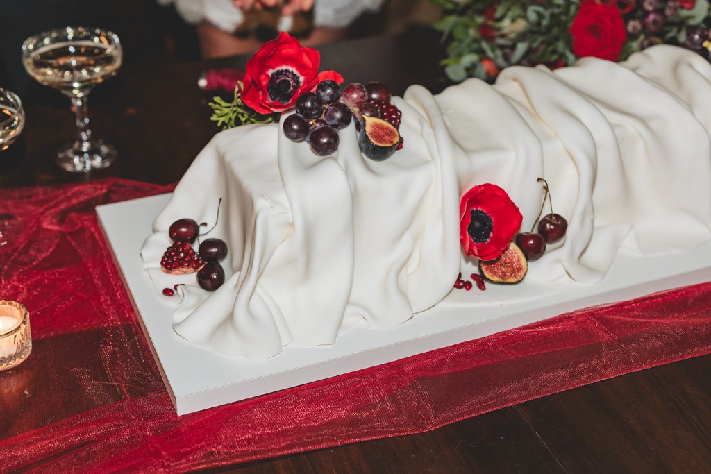 A contemporary, design-led wedding cake styled with seasonal fruit and striking red anemone accents.