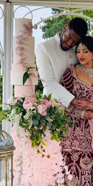 Design-led wedding cake featuring peach and blush tones, sculptural tiers and cascading florals, thoughtfully styled within a contemporary outdoor pagoda wedding setting in Northamptonshire, alongside the bride and groom